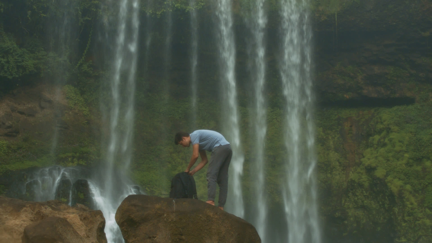 Man tourist takes smartphone from backpack standing on huge rock and makes selfie against fantastic waterfall streams