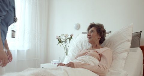Home Nurse Making Bed of Elderly Female Patient. Female Caregiver Adjusting Pillow For Senior Woman Lying in Hospital Bed.
