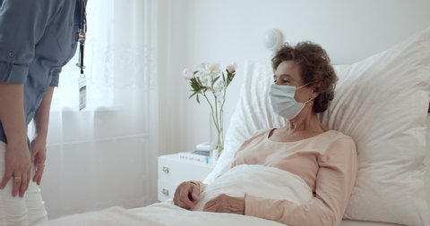 Elderly Woman Wearing Protection Face Mask Having Friendly Communication With Nurse in Hospital Room. Home Caregiver With Face Mask Holding Hands of Female Senior Patient Lying in Bed at Nursing Home.