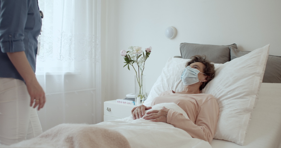 Home Caregiver With Face Mask Comforting Female Senior Patient Lying in Bed at Nursing Home. Elderly Woman Wearing Protection Face Mask Reaching Out To Nurse Approaching Her Hospital Bed. Royalty-Free Stock Footage #1061436220