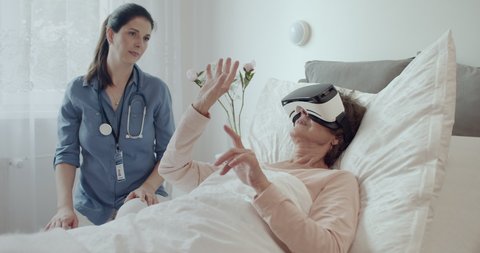 Female Doctor Checking on Elderly Patient Lying in Hospital Bed Doing Therapy Via VR Technology. Friendly Doctor Helping Senior Woman Receive Therapy Using Virtual Reality Headset.