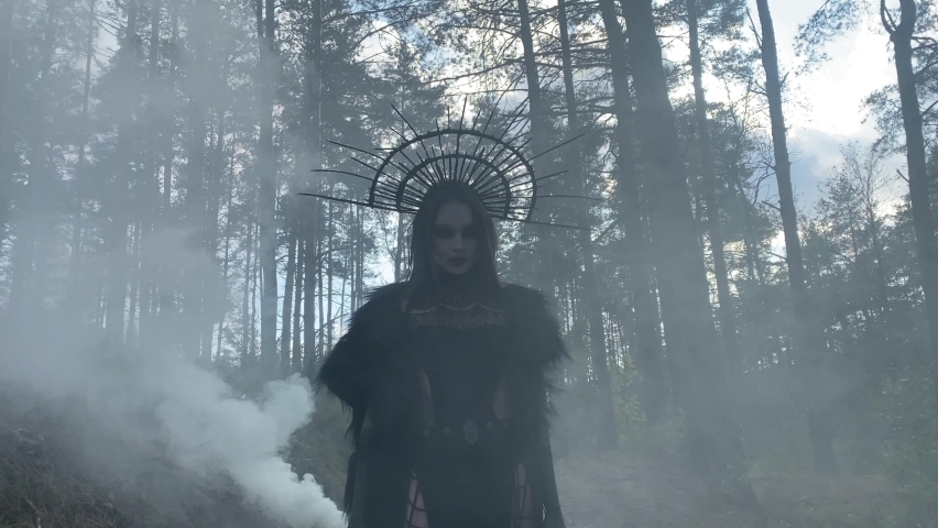 Young woman in image of witch walks in black dress and crown on her head in penetrated by the sun rays foggy forest.