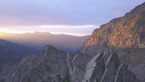 Rugged mountain peaks and narrow sharp ridge high in mountains of Romania with stunning sunset sky filled with purple and red. Glow of setting sun shines on cliff face. Aerial view from drone.
