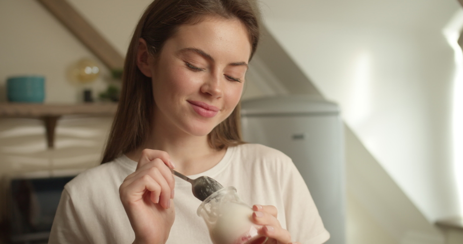 Incredible girl brunette of Slavic appearance in a white T-shirt holds a metal spoon and a glass jar with yogurt, which eats, enjoying every moment, transferring the look into the camera. | Shutterstock HD Video #1061444305