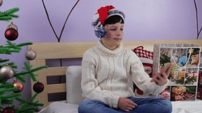 Christmas online greetings. A smiling boy in a white knitted sweater with a gift uses a mobile phone for video calls with friends and parents. Shot in 4k