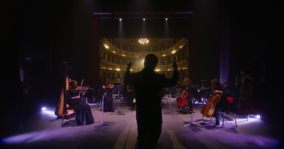 Cinematic shot of conductor directing symphony orchestra with performers playing violins, cello and trumpet on classic theatre with curtain stage during music concert with dramatic lights. Royalty-Free Stock Footage #1061452294