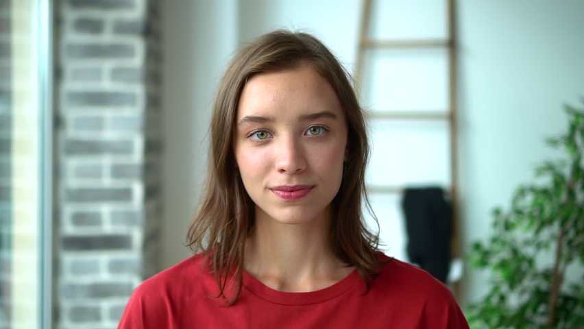 Portrait of beautiful young woman posing in front of camera while standing in light interior avki. Face view of American young woman looks forward with toothy smile and poses at room with daylight