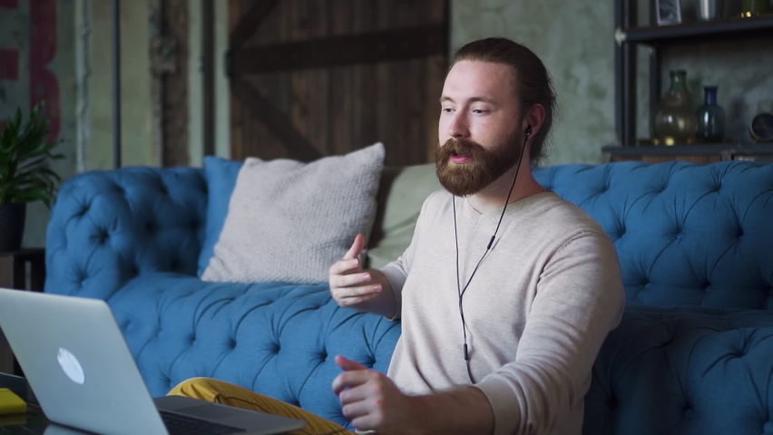 Young bearded businessman discusses business and has online call on couch in apartment spbd. Handsome man is talking and gesturing while looking at computer screen in bright room. One red-headed guy
