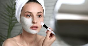 Healthy young woman with pure skin using brush for apllying mask on face while sitting at bathroom and looking at mirror. Spa and beauty at home.
