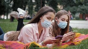 Young women in medical masks lie in the park on the grass and using smartphone or cell phone. Tapping, scrolling, watching video, content, blogs.