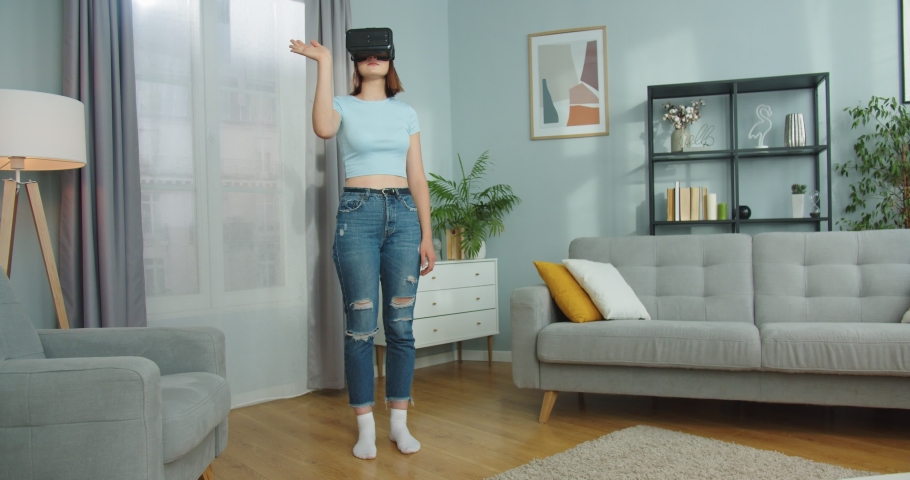 Beautiful Caucasian female using vr glasses, exploring virtual reality in living room. Inquisitive young woman in futuristic googles scrolling web sites on Internet. Technology, future concept. | Shutterstock HD Video #1061467456