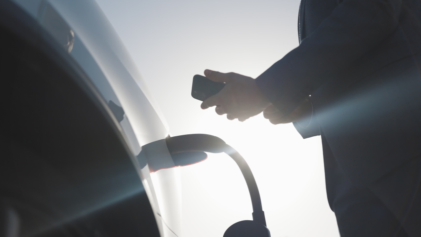 Unrecognizable businessman plugging electric car from charging station. Senior is plugging in power cord to an electric car at sunset. Caucasian businessman charging electric car at charging station | Shutterstock HD Video #1061468395