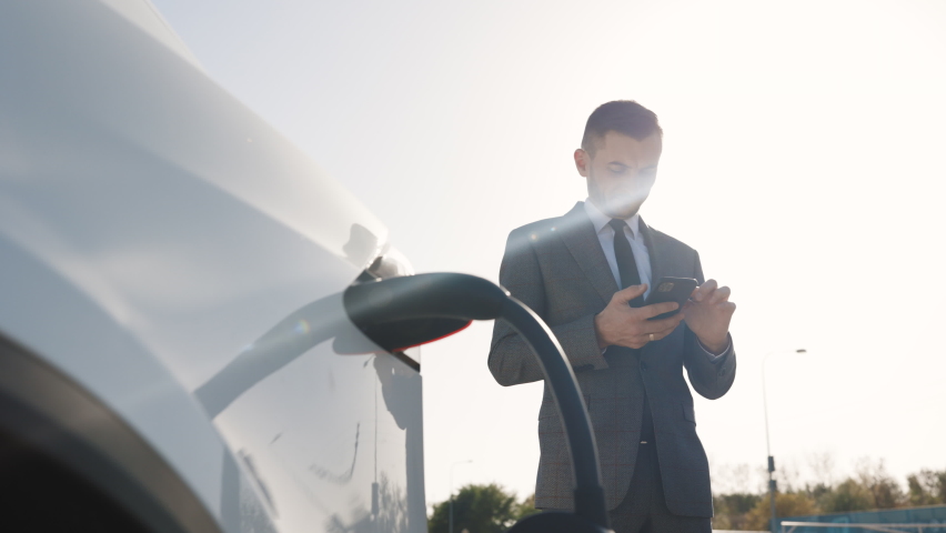 Caucasian businessman using smart phone and waiting power supply connect to electric vehicles for charging the battery in car. Plug charging an Electric car from charging station. Royalty-Free Stock Footage #1061468404
