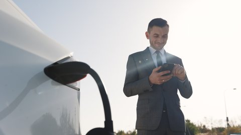 Caucasian businessman using smart phone and waiting power supply connect to electric vehicles for charging the battery in car. Plug charging an Electric car from charging station.