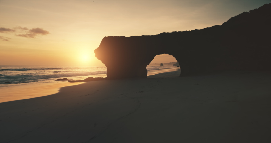 Slow motion sun silhouette of rock wall with giant hole on sand beach aerial view. Sandy ocean coast, unique cliff at Sumba Island, Indonesia, Asia. Nobody nature at sunlight. Cinematic summer sunset
