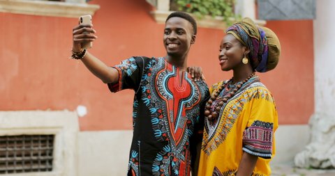African young happy and good looking man and woman in traditional clothes standing together outdoor and taking selfie photos on the smartphone camera.