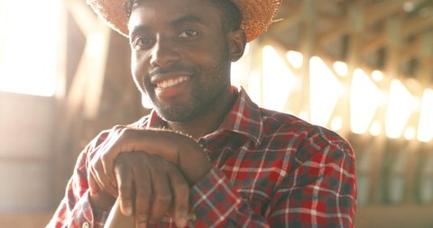 Portrait of young handsome African American man farmer in hat smiling at camera and leaning on pitchfork in barn. Good-looking cheerful male shepherd smile in dark stable. Close up. Flies flying.