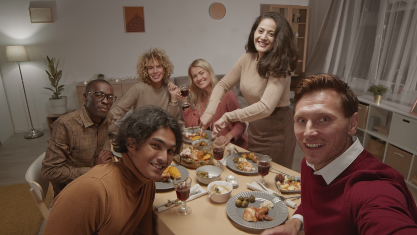 Medium shot of young Caucasian man holding camera in his hands and making selfie with his diverse friends sitting at dining table, looking at camera and waving hands Royalty-Free Stock Footage #1061475499