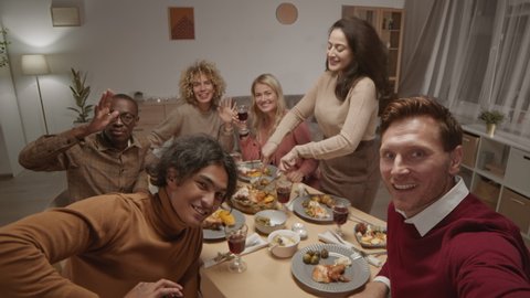 Medium shot of young Caucasian man holding camera in his hands and making selfie with his diverse friends sitting at dining table, looking at camera and waving hands
