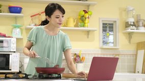 attractive asian woman cooking while watching laptop in the kitchen