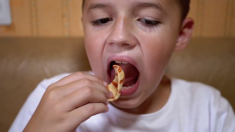 Hungry Caucasian Child Puts Chips in Mouth with Hand. Boy Eats Fast Food. Snacking on junk food. Portrait of a boy with a pretty face. Tasty food. Fast and unhealthy food concept. Obesity. 180fps