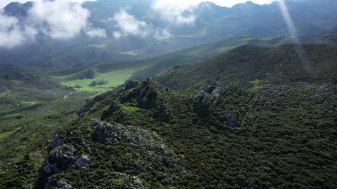 Aerial view overlooking a valley and a road, between green mountains and low clouds, in the Peaks of Europe National Park, sunny summer day, in Asturias, Spain - tracking, pan shot