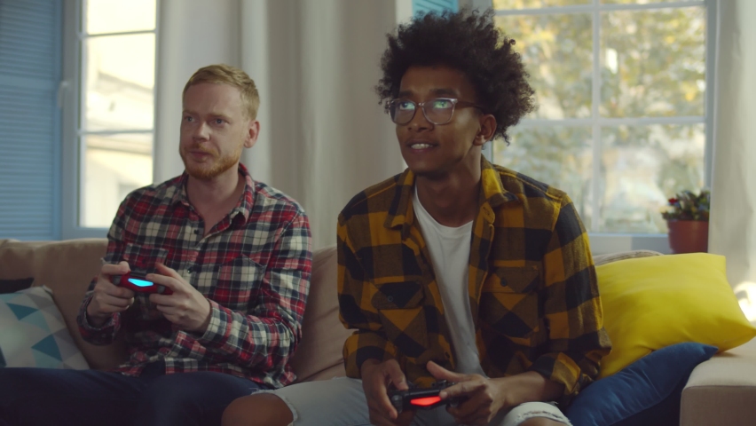 Smiling diverse male friends with gamepads playing video game at home. Portrait of african and caucasian hipster guys playing game with console relaxing on couch in living room Royalty-Free Stock Footage #1061492281