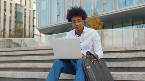 Young african businessman using laptop while sitting on the stairs outdoors. Afro american student studying on computer sitting on campus staircase outside. Entrepreneur working on laptop