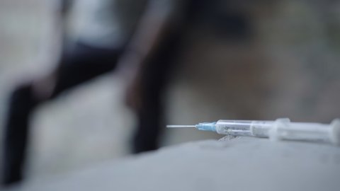 Close-up of syringe with blurred junkie writhing at the background. Unrecognizable overdosed drug addict on urban ruins in ghetto. Drug trafficking and substance addiction.