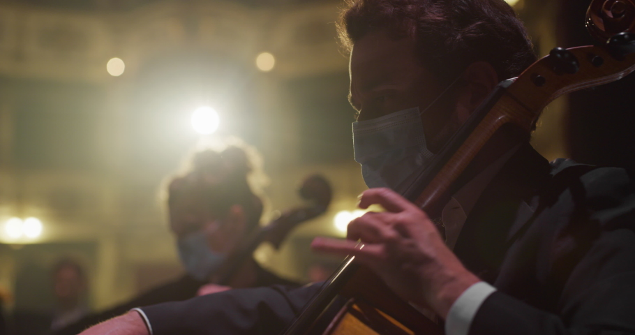 Cinematic Close Up Shot of Symphony Orchestra Cello Player Wearing Medical Protective Mask on Classic Theatre with Curtain Stage during Music Concert. Performers Playing Music for Audience