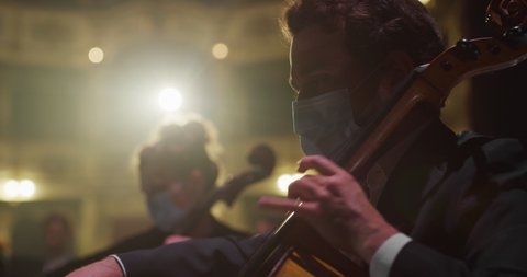 Cinematic close up shot of symphony orchestra with performers wearing medical protective masks playing violins, cello and trumpet on classic theatre with curtain stage during music concert.
