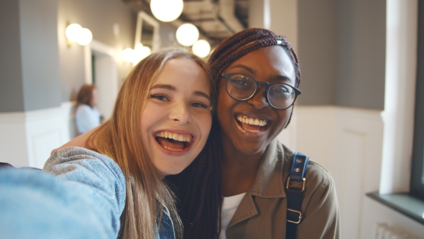 International female students with beaming smile posing for selfie shot. Portrait of two diverse young women bloggers filming video for internet blog indoors Royalty-Free Stock Footage #1061494300