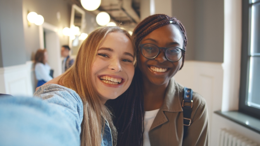 International female students with beaming smile posing for selfie shot. Portrait of two diverse young women bloggers filming video for internet blog indoors