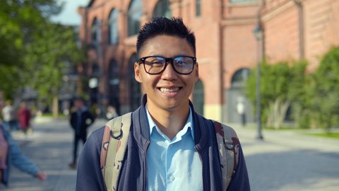 Portrait of young asian man in eyeglasses smiling and looking at camera. Attractive chinese male college student with backpack in campus. Happy guy standing outdoors over red brick building background