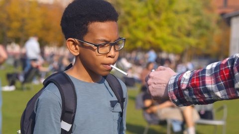 Portrait of african preteen boy taking cigarette from dealer and smoking. Hand with lighter lighting cigarette for underage afro-american schoolboy in park