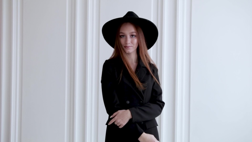 elegant girl in a dress and a black hat smiling and posing in a white room with large Windows, slow motion