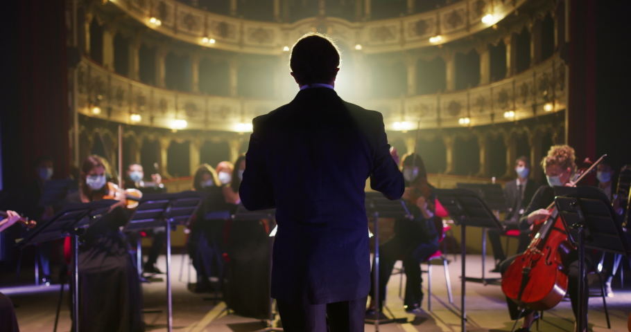 Back View of Professional Conductor Directing Symphony Orchestra with Performers Wearing Medical Masks, Playing Violins, Cello and Trumpet on Classic Theatre with Curtain Stage During Music Concert Royalty-Free Stock Footage #1061499835
