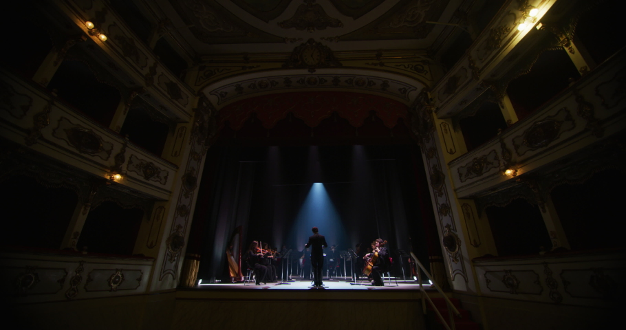 Wide shot of an Orchestra on a Classic Theatre Stage:  Professional Conductor Directing Symphony Orchestra with Performers Playing Violins, Cellos, and Trumpets During Music Concert. Audience's POV Royalty-Free Stock Footage #1061499865