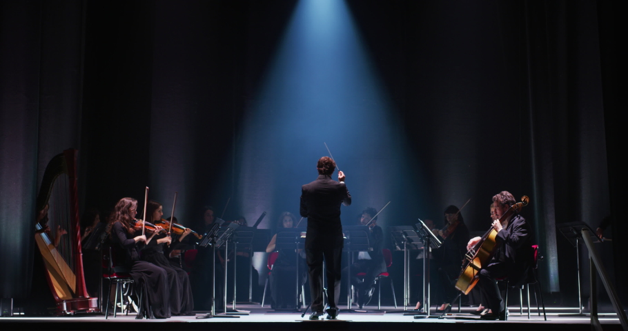 Wide shot of an Orchestra on a Classic Theatre Stage:  Professional Conductor Directing Symphony Orchestra with Performers Playing Violins, Cellos, and Trumpets During Music Concert. Audience's POV Royalty-Free Stock Footage #1061499877