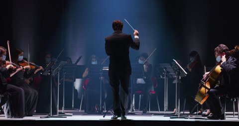 Cinematic shot of conductor directing symphony orchestra with performers with medical protective masks playing violins, cello and trumpet on classic theatre with curtain stage during music concert.