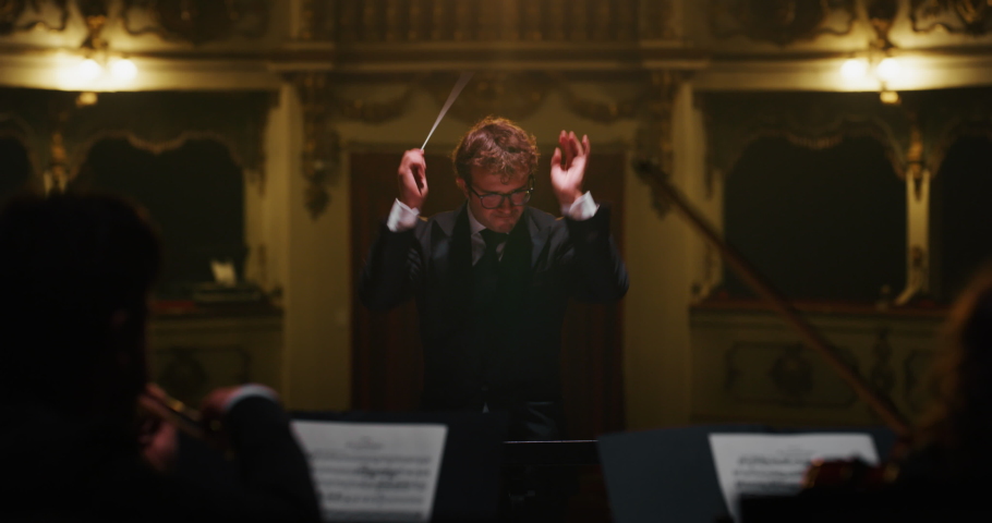 Cinematic Close Up of Conductor Directing Symphony Orchestra with Performers Playing on Stage During Music Concert. Professional Conductor Leading Musicians Passionately in Classic Theater Royalty-Free Stock Footage #1061500405