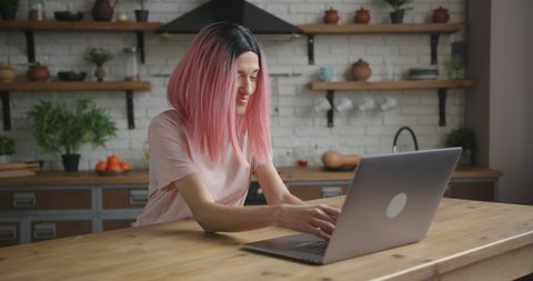 Pretty elegant transwoman with stylish pink wig works on modern laptop sitting at wooden table in decorated kitchen at home