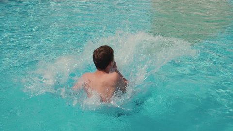 Boy relaxing during summer vacation, slow motion. Unrecognizable guy jumping in pool and splashing crystal clear water in basin at sunny day.