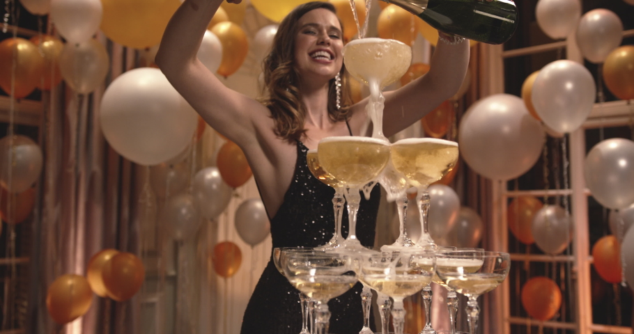 Beautiful young woman pouring two bottles of champagne into glasses tower at gala night party. Smiling female in evening gown filling pyramid of glasses with champagne.
 | Shutterstock HD Video #1061502139
