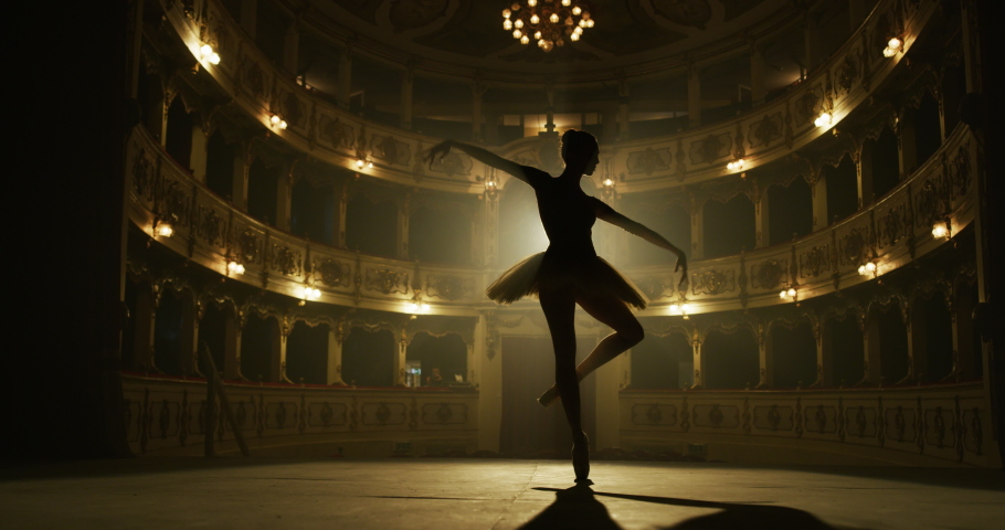 Cinematic shot of an young graceful classical ballet female dancer in white tutu is performing a choreography on classic theatre stage with dramatic lighting before start of a show.