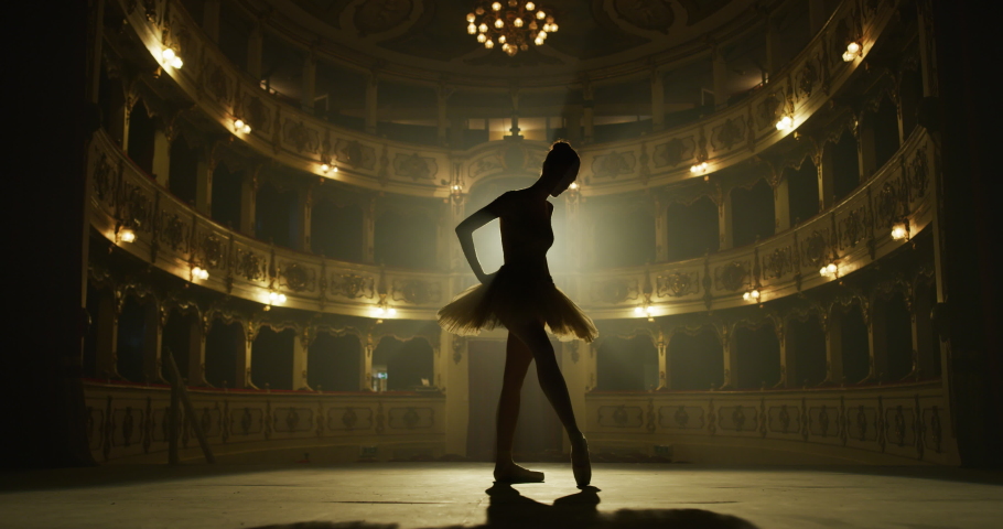 Cinematic shot of an young graceful classical ballet female dancer in white tutu is warming up for performing a choreography on classic theatre stage with dramatic lighting before start of a show.