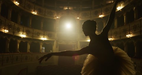 Cinematic close up shot of an young graceful classical ballet female dancer in white tutu is performing a choreography on classic theatre stage with dramatic lighting before start of a show.