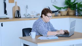 Senior woman using laptop for websurfing in her kitchen. The concept of senior employment, social security. Mature lady sitting at work typing a notebook computer in an home office. Shot video