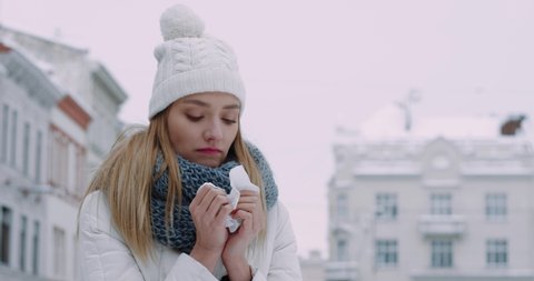 Young beautiful woman in winter clothes feels sick and exhausted blowing her nose into handkerchief while standing outside in bad winter weather.