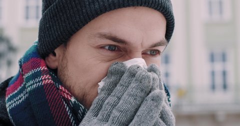 Young Caucasian man in winter clothes standing on the street and blows his nose into handkerchief. Symptoms of cold in winter.